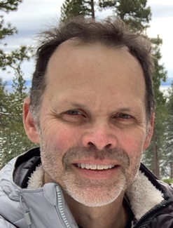 Professor Lloyd Knox poses outdoors wearing a puffy jacket in front of a winter landscape with tall trees.